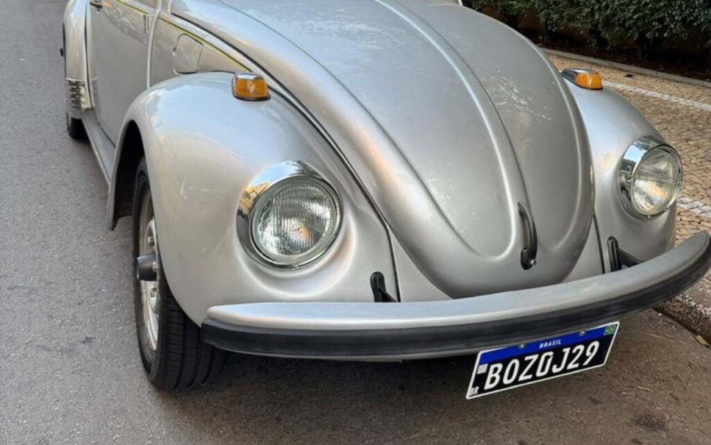 Fusca prateado antigo com placa preta do Brasil estacionado em rua asfaltada, com faróis arredondados, parachoque robusto e design característico dos carros clássicos.
