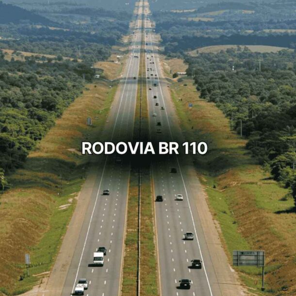 Fotografia aérea da Rodovia BR 110 com pista dupla, vários veículos circulando em ambos os sentidos, cercada por áreas verdes, vegetação densa e morros ao fundo, destacando o tráfego intenso na Rodovia BR 110.