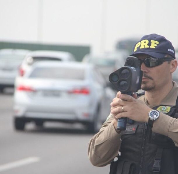 Policial rodoviário federal usa radar portátil para fiscalizar velocidade em rodovia movimentada, com vários carros ao fundo, representando fiscalização que pode gerar multa RENAINF por infração em outro estado.