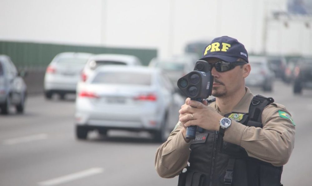 Policial rodoviário federal usa radar portátil para fiscalizar velocidade em rodovia movimentada, com vários carros ao fundo, representando fiscalização que pode gerar multa RENAINF por infração em outro estado.