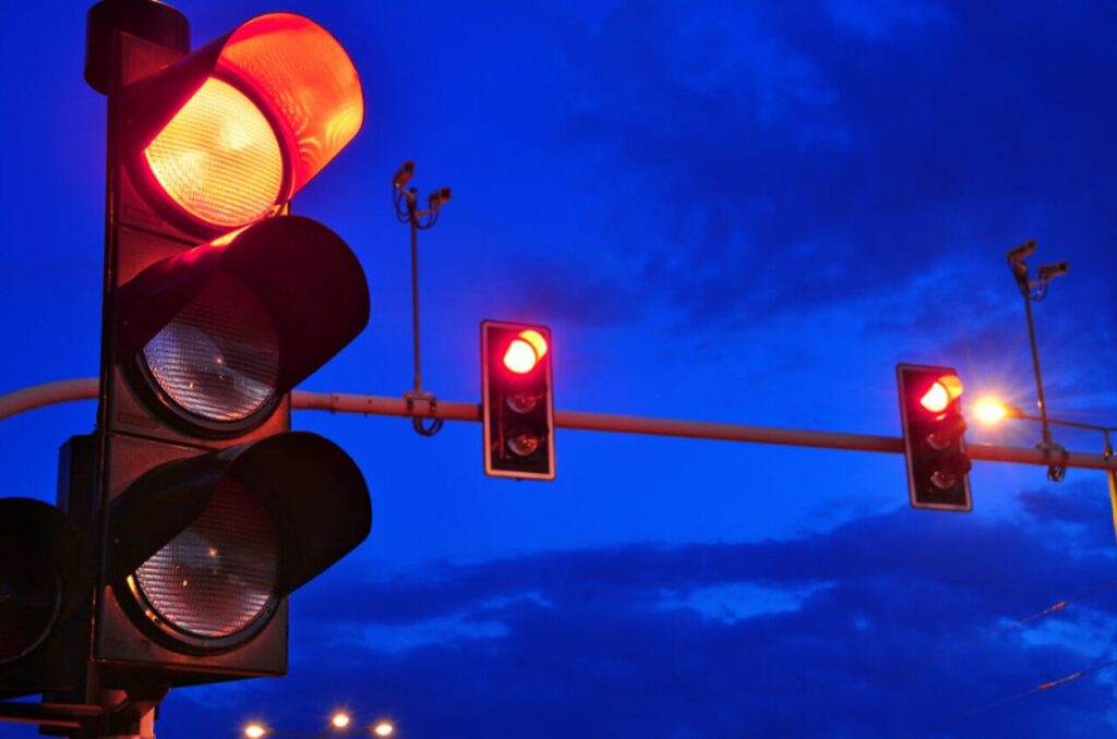 Foto de vários semáforos com luz vermelha acesa ao entardecer, sob um céu azul escuro. A imagem ilustra o risco de multa por passar no sinal vermelho, especialmente em horários de menor visibilidade.