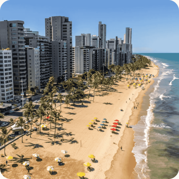 Praia em Recife-PE com faixa de areia dourada, guarda-sóis coloridos e altos edifícios residenciais ao fundo sob céu azul — turismo e infraestrutura costeira em destaque para licenciamento 2025.