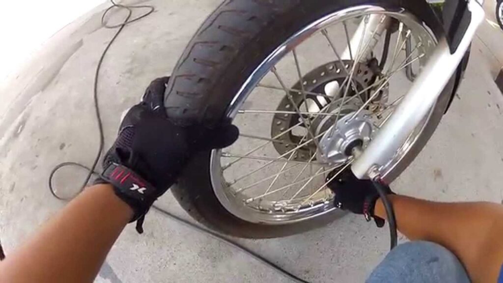 Pessoa usando luvas preta inflando o pneu dianteiro de uma moto com calibrador, em ação prática de manutenção preventiva com foco na calibragem do pneu da moto.
