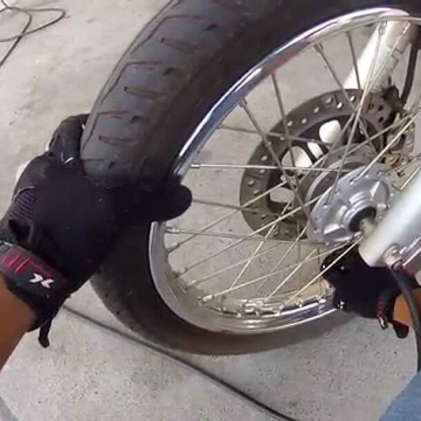Pessoa usando luvas preta inflando o pneu dianteiro de uma moto com calibrador, em ação prática de manutenção preventiva com foco na calibragem do pneu da moto.