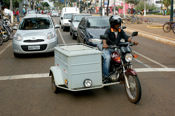 Moto circulando no trânsito urbano com carretinha lateral acoplada, utilizada para transporte de mercadorias, exemplo comum de carretinha lateral para moto em áreas comerciais.