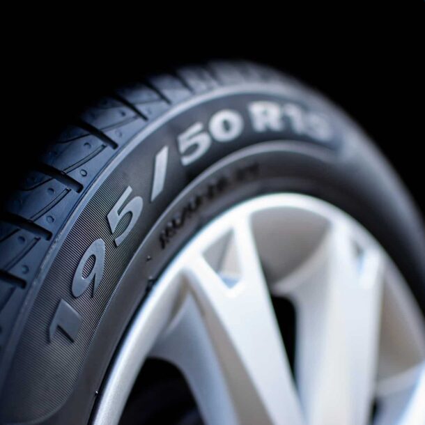 Close em pneu de carro com a numeração do pneu 195/50 R15 visível na lateral, destacando detalhes da banda de rodagem e o aro prateado da roda em fundo escuro.