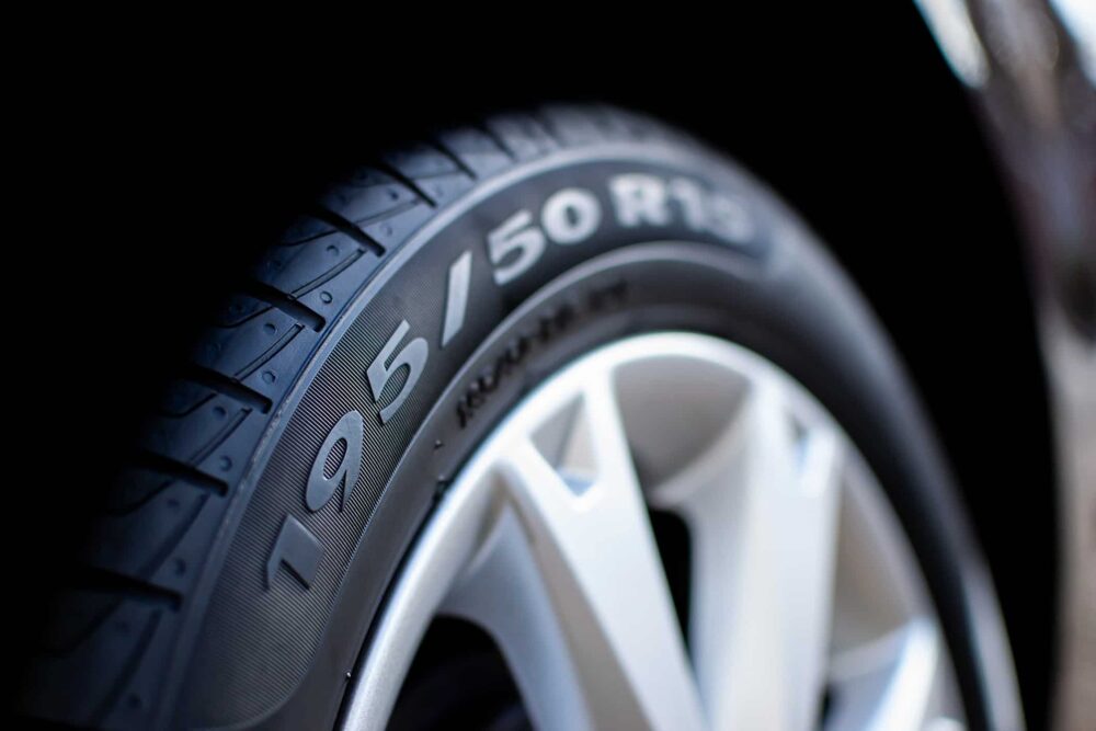 Close em pneu de carro com a numeração do pneu 195/50 R15 visível na lateral, destacando detalhes da banda de rodagem e o aro prateado da roda em fundo escuro.