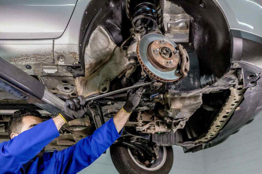 Mecânico com luvas e uniforme azul realiza manutenção na suspensão automotiva de um carro elevado, ajustando componentes da roda e do sistema de freio com chave de força.