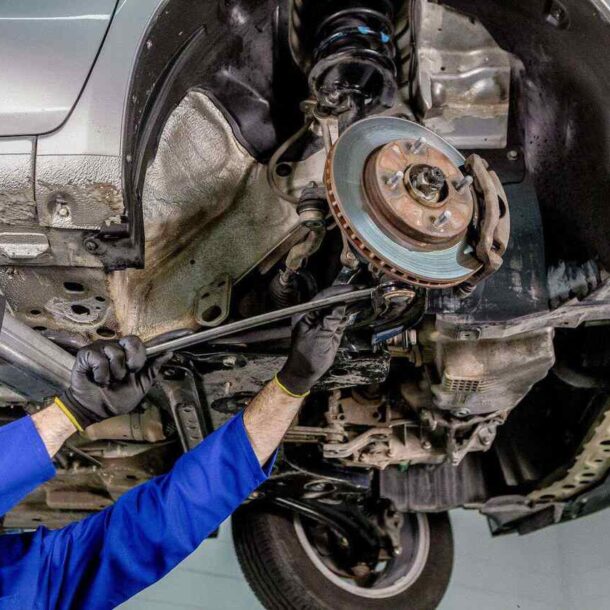 Mecânico com luvas e uniforme azul realiza manutenção na suspensão automotiva de um carro elevado, ajustando componentes da roda e do sistema de freio com chave de força.
