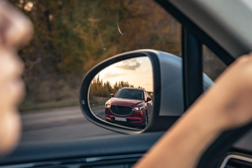 Retrovisor de carro mostra veículos vindo atrás ao pôr do sol em estrada. A composição reforça a importância dos espelhos e a possibilidade de multa por andar sem retrovisor em vias públicas.