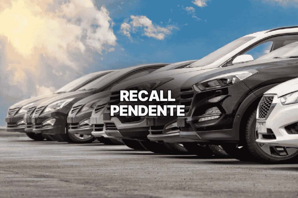 Imagem de vários carros estacionados em fila sob céu azul com nuvens, acompanhada do texto “recall pendente” em destaque central, alertando sobre a necessidade de verificar pendências veiculares.
