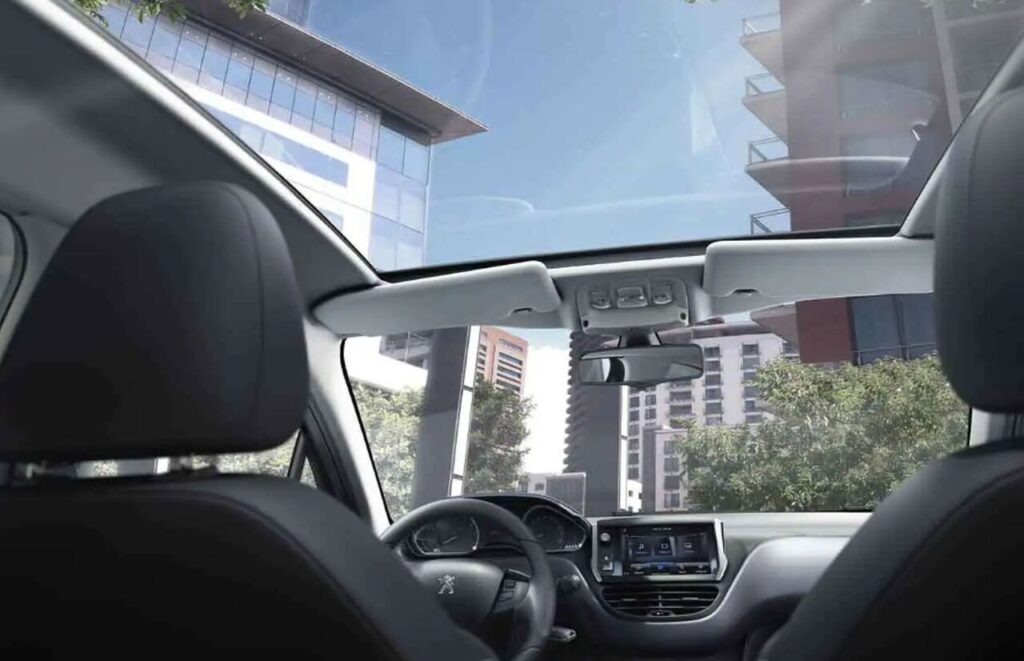 Interior de carro com teto solar panorâmico aberto, mostrando visão ampla de prédios modernos e céu azul através do para-brisa e do vidro superior