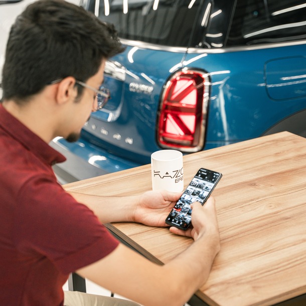 Motorista de aplicativo compara carros pelo celular em uma concessionária antes de financiar o veículo.
