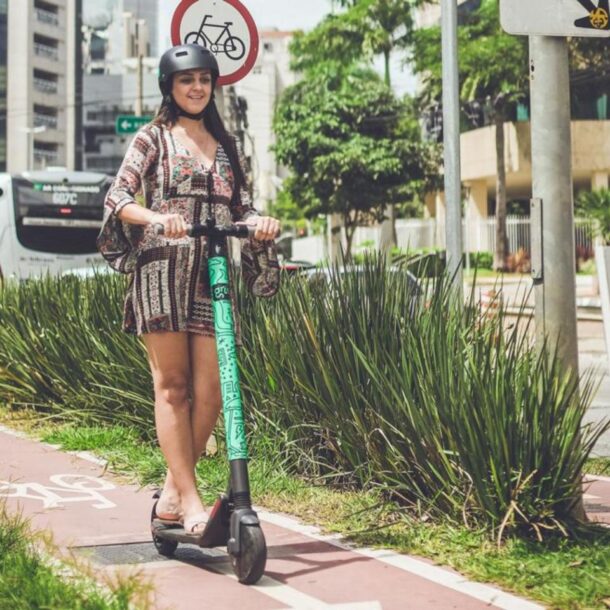 Mulher conduz patinete elétrico em ciclovia urbana usando capacete.
