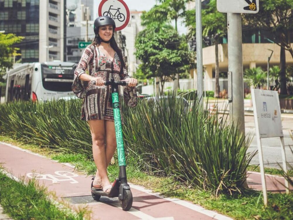 Mulher conduz patinete elétrico em ciclovia urbana usando capacete.
